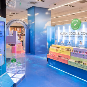 A brightly lit cosmetics store with colorful displays and a modern design. Custom retail displays showcase a variety of beauty products in pastel hues. A large sign reads CLEAN, COOL & COVETED, echoing the Sephora Clean ethos. The floor has a blue, cloudy pattern.