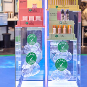 A vibrant Sephora Retail Display showcases colorful cosmetic products on clear stands, adorned with green check marks. Featuring makeup essentials like lipsticks and foundations in diverse packaging, it stands out against a bright retail environment backdrop.