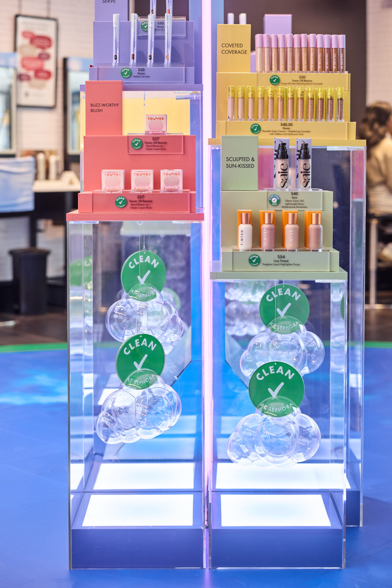 A vibrant Sephora Retail Display showcases colorful cosmetic products on clear stands, adorned with green check marks. Featuring makeup essentials like lipsticks and foundations in diverse packaging, it stands out against a bright retail environment backdrop.