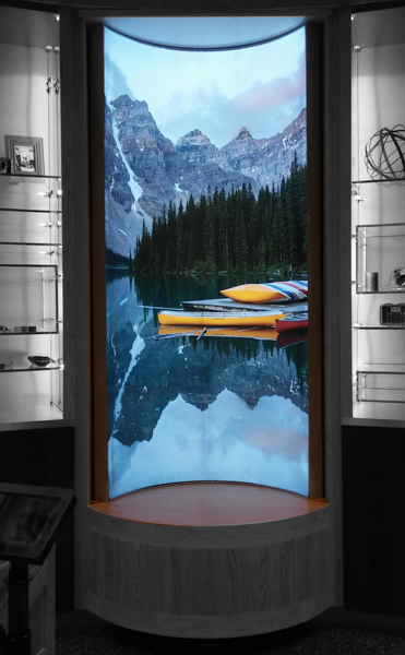 A curved display showcases a serene mountain lake scene with canoes on the water and pine trees reflecting in the lake, framed by shelves adorned with artifacts like small sculptures and books.