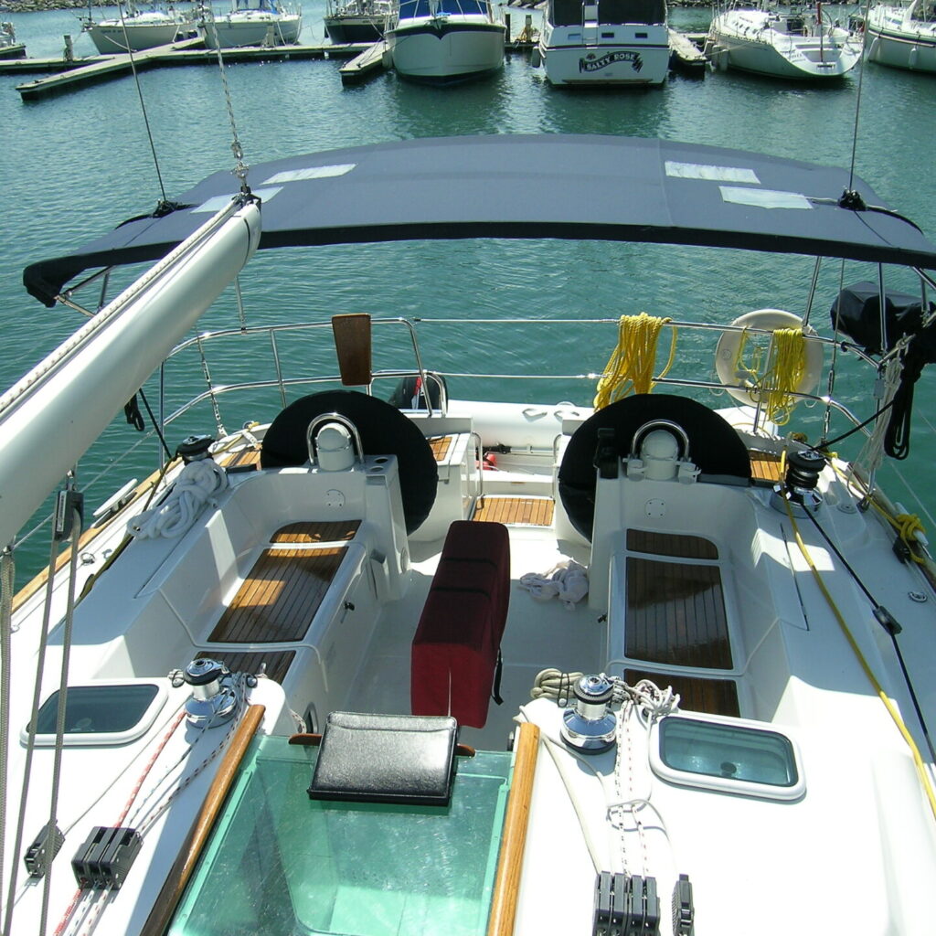 POV From the helm of a sailboat docked in the marina. The shaded cockpit boasts two steering wheels, seating, and a red cushion. Other boats and yachts shimmer in the background under the sunny sky.