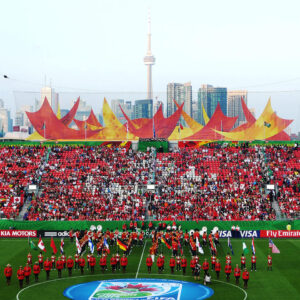 A lively stadium scene unfolds as a marching band performs on the field. The stands are packed with fans in red, cheering for the FIFA Women's Cup. Toronto's skyline, including the CN Tower, towers in the background as a giant banner is held up by cranes, adding to the colorful atmosphere.