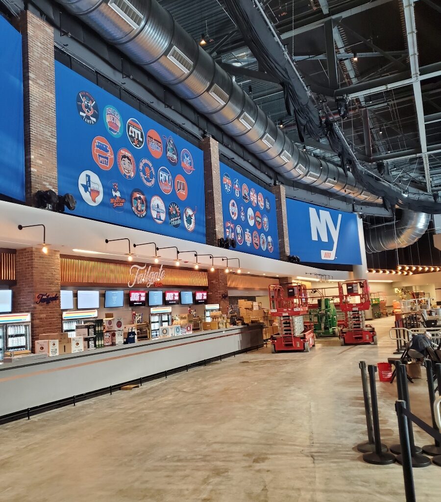 A spacious modern interior with a long bar counter on the left. Above the counter, large blue displays showcase various logos with NY prominently featured. The ceiling's exposed pipes and ducts add an industrial charm, while construction equipment occupies the area.