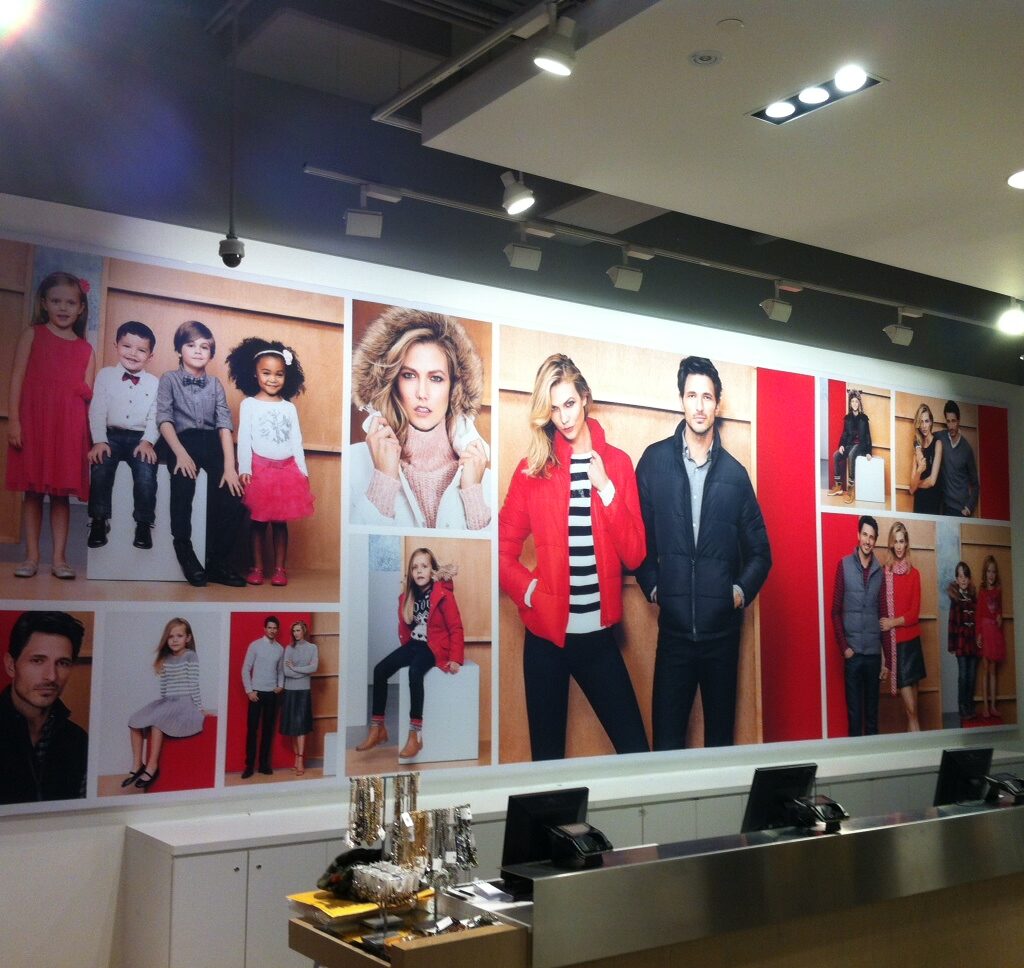 A large Joe Fresh SEG Fabric Display features various fashion images, including adults and children modeling jackets, dresses, and casual wear. The setting includes a checkout counter with lights overhead in a vibrant retail environment.
