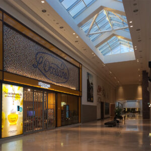 Modern shopping mall corridor with a high arched glass ceiling, empty except for benches and plants. A LOccitane storefront with an elegant retail display is illuminated on the left, showcasing products and a vibrant poster. The brightly lit interior feels expansive and inviting.