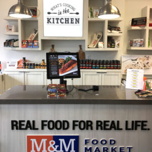Interior of an M&M Food Market store with a counter displaying products, a screen, and a lightbox sign above reading "What's Cooking in the Kitchen.