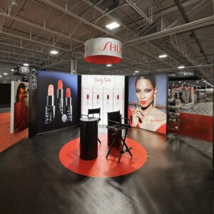 A makeup exhibition booth featuring lipstick displays and promotional images. There are two black chairs in the center on a circular red platform, with brand names like NARS visible on the banners. Overhead, a circular sign hangs from the ceiling.