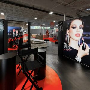 Cosmetic store display featuring a NARS advertisement with a model holding a lipstick. The setup includes a black chair and cylindrical stands, surrounded by bright red accents. The background shows a spacious, well-lit Trade-show interior.