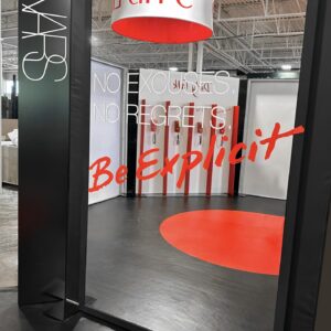 A retail display with bold black and red decor. A large mirror displays the words NO EXCUSES. NO REGRETS. Be Explicit in red. The NARS logo appears on the frame. Red and white cylindrical light fixtures hang from the ceiling.