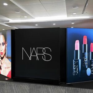 NARS store display featuring a large black sign with the brands logo. On the left, an image of a model with makeup. On the right, colorful lipsticks and lipstick tubes against a blue background.