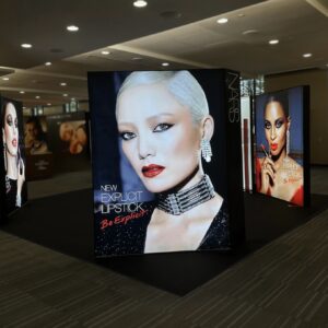 A modern makeup display featuring a model with short white hair, wearing dramatic eye makeup and bright red lipstick. The background advertises NARS New Explicit Lipstick: Be Explicit. Another similar display is partially visible in the background.