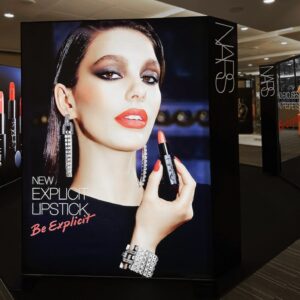 A makeup advertisement showcases a model holding a red lipstick, wearing bold makeup and dangling earrings. Text reads NEW explicit lipstick - Be Explicit with the brand logo NARS prominently at the top right.