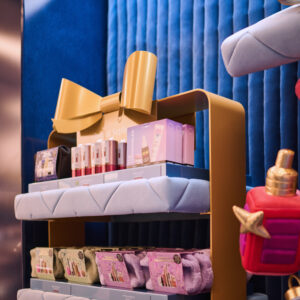 Cosmetics display in a store with various beauty products arranged on a shelf. The shelving unit has a large gold bow on top and is adorned with plush-looking purple material. The background features textured blue fabric.