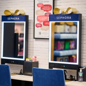A beauty counter with two mirrors, each beneath a sign labeled Sephora. The mirrors are surrounded by soft white lights. Blue fabric covers two chairs at the counter, which is stocked with beauty supplies and sanitizer.