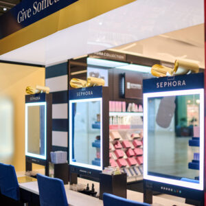 A Sephora store display featuring three makeup stations with illuminated mirrors and blue chairs. Shelves in the background hold various beauty products. The stores name is visible above the mirrors.