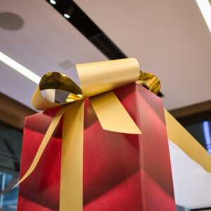 A large, decorative gift box wrapped in red paper with a shiny gold ribbon and bow. The box is placed indoors, and the ceiling with lights is visible in the background.