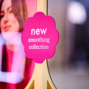 A pink flower-shaped sign reads “new smoothing collection” in white text, placed in front of a blurred image of a woman with long hair wearing a pink top.