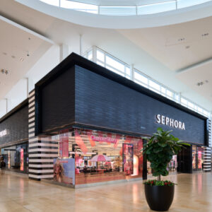 A Sephora store with large glass windows displaying beauty products, located inside a modern, brightly lit shopping mall with beige tile flooring and a potted plant in the foreground.